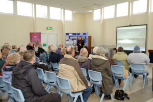 A photo showing a room full of people listening to a person speak in front of a large projector screen.